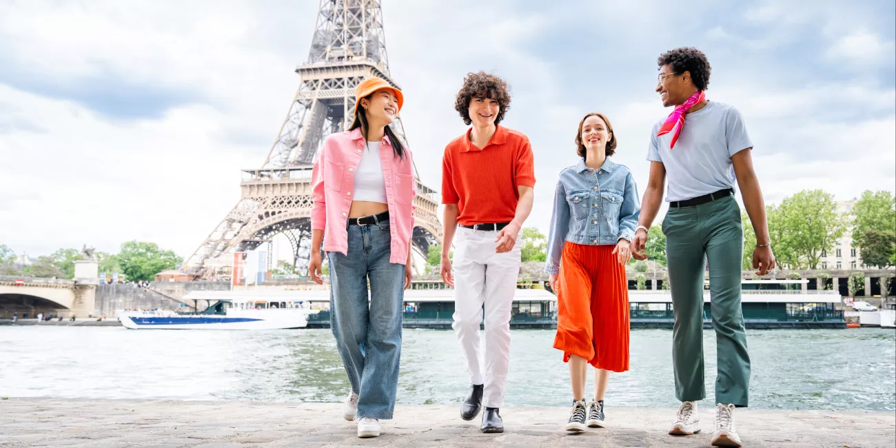 Four young people walk together along the Seine with the Eiffel Tower in the background, smiling and chatting on a bright day.