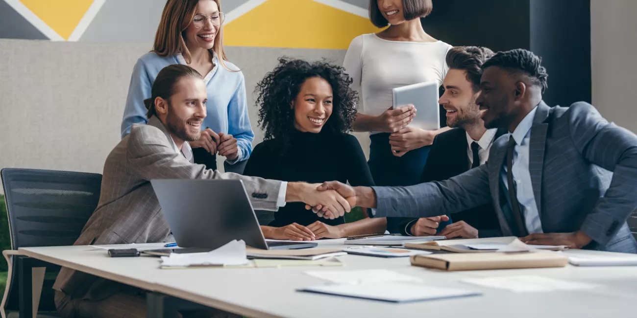 Two smiling young businessmen shaking hands while sitting with their colleagues at the office desk