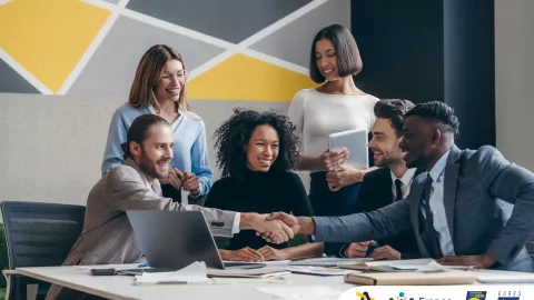 Two smiling young businessmen shaking hands while sitting with their colleagues at the office desk