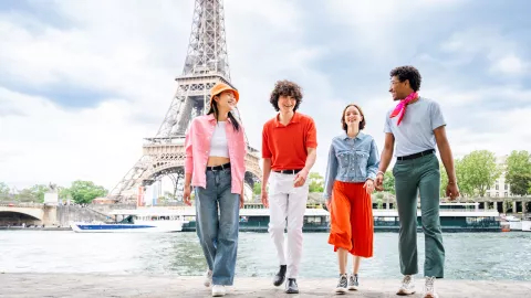 Four young people walk together along the Seine with the Eiffel Tower in the background, smiling and chatting on a bright day.