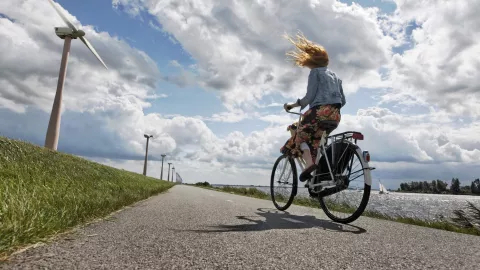 young woman on her bicycle traveling on a wind blown dike in Holland, wind turbines in the background